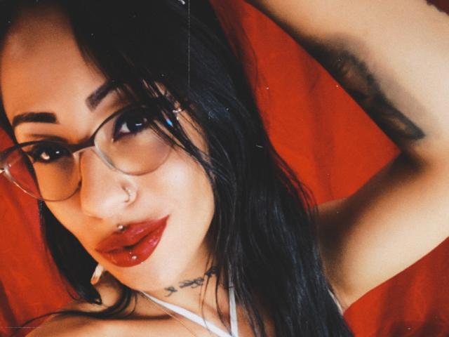 LunaCandy met een bril, zwart haar, rode lipstick, een neusring en een lipring, draagt een rode top, heeft haar armen omhoog en laat een tatoeage op haar oksel zien. Haar borsten zijn klein, ze is schattig..
