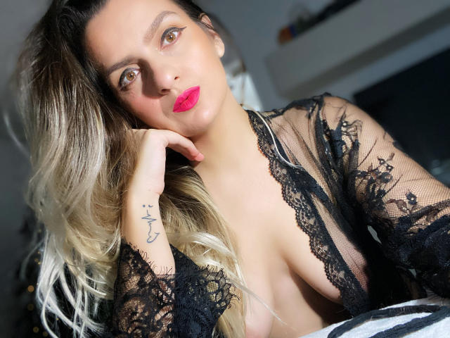ReveMignoneX with blonde, wavy hair, wearing black lace, exposing her tits, has pink lipstick, tattoo on left hand, and a sexy, confident look in a bright room.