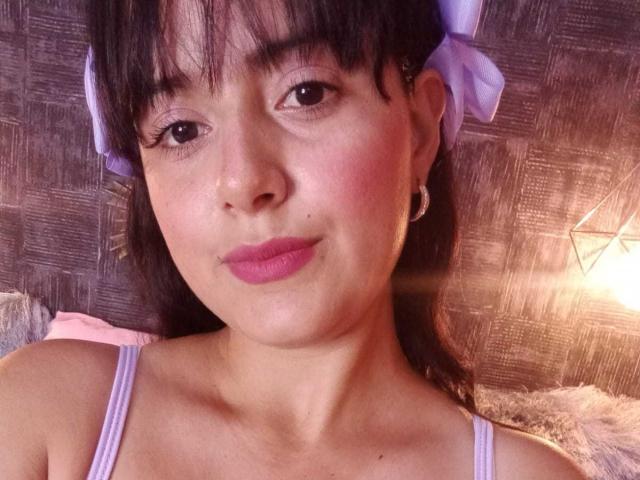 LanaLorens has pink lipstick, purple ribbons in her black hair, and a cute smile, wearing a lavender top, with subtle blush on her fair skin.