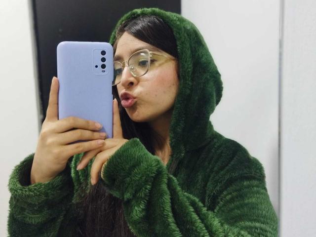EvaMcAllister, com um capuz verde felpudo, lábios curvados, segurando um telefone azul, usando óculos de aro bege, tem cabelos longos e escuros, pele clara, em um selfie..