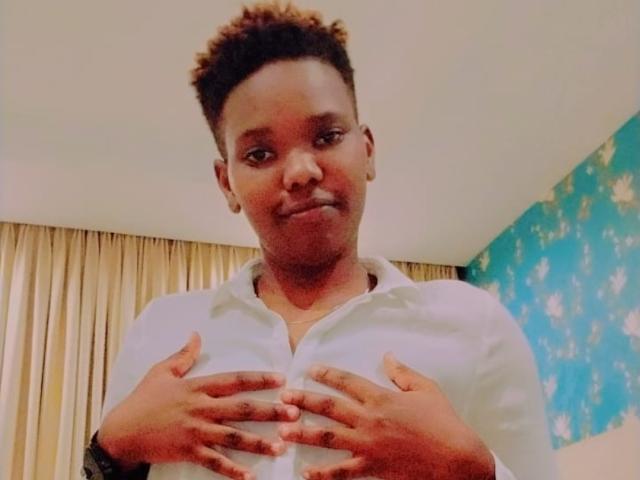 SandyHottLove, a dark-skinned person with short curly hair, is smiling and holding their tits with hands, wearing a white button-up shirt.
