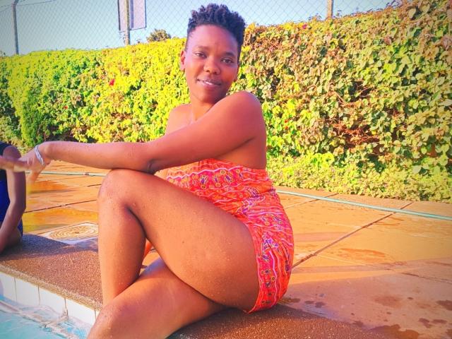 SandyHottLove, a dark-skinned woman with short curly hair, sits by the pool in a red, patterned one-piece, showing her ass, with a smile, arms crossed.