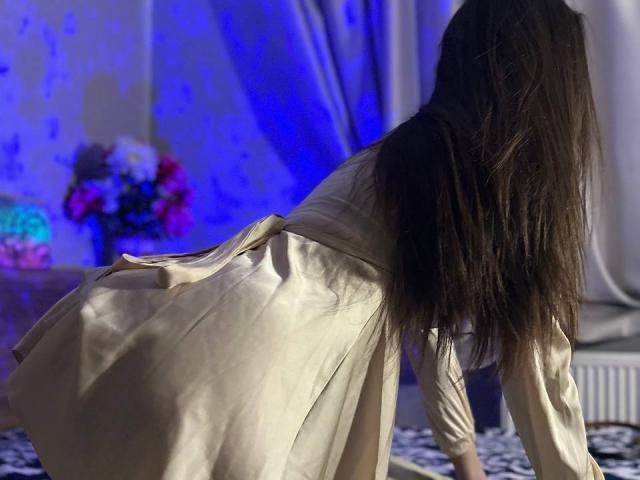 CheriLady, con lunghi capelli castani, indossa un abito beige di seta ed è in posizione a quattro zampe, con glutei e schiena visibili, in una stanza poco illuminata con luci blu..