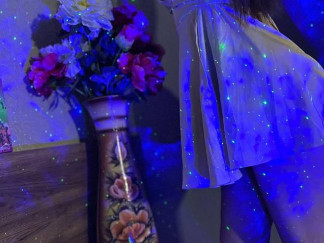 CheriLady's white, sheer, sparkly dress with blue lights, exposing her ass, standing beside a vase with colorful flowers. Her dress is translucent, showing her skin.