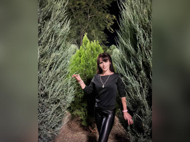BeatriceHottLove si trova in una foresta oscura, indossa una maglietta nera, pantaloni di pelle nera lucida e una collana d'oro, con la mano destra sollevata, circondata da cespugli verdi..