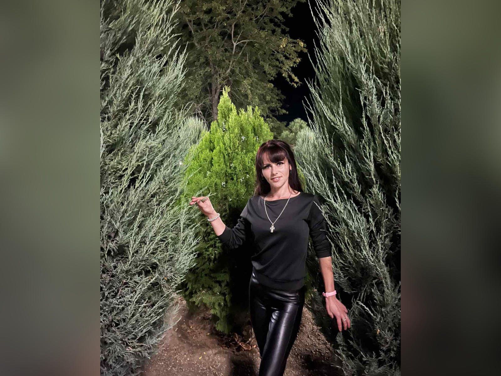 BeatriceHottLove está de pie en un bosque oscuro, vestida con una camisa negra, pantalones de cuero negro brillante y un collar dorado, con la mano derecha levantada, rodeada de arbustos verdes..