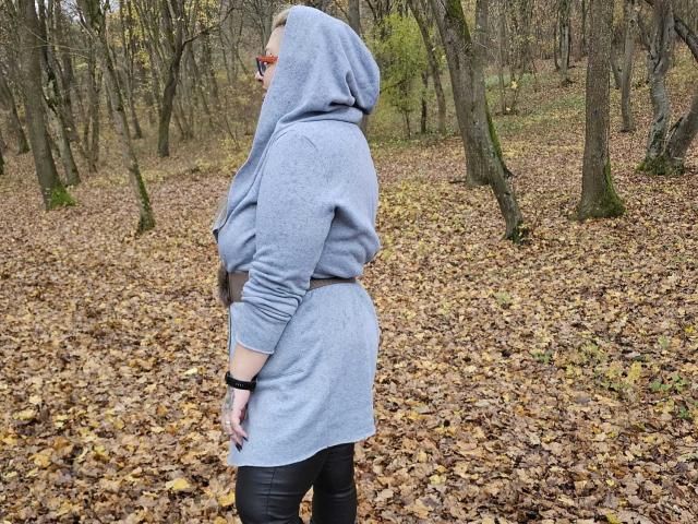 BustyYola in a gray hooded sweater, standing sideways, showing her ass, wearing black pants, in a forest with a leaf-covered ground. She's looking over her shoulder.