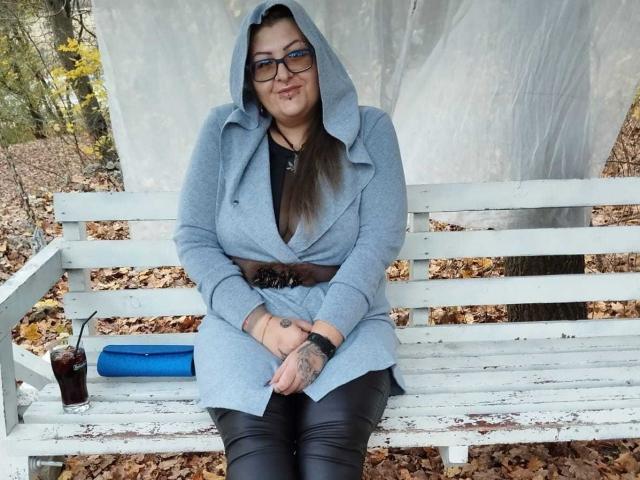 Yola, avec une poitrine généreuse, portant des lunettes, un sweat à capuche gris, un pantalon noir, assise sur un banc blanc, les mains posées sur ses genoux, une boisson noire avec une paille sur la gauche, un objet bleu sur la droite, des feuilles d'automne, un tatouage au poignet..
