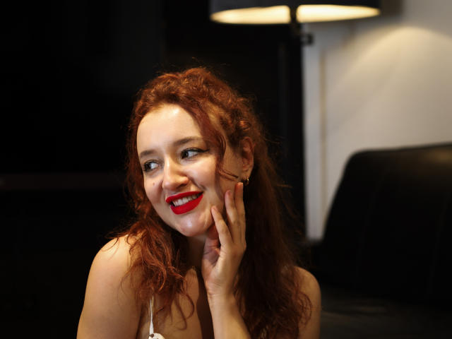 LailaNoire with curly red hair, red lipstick, smiling, touching her cheek, wearing a white top, in a dimly lit room with a black lamp and leather chair.