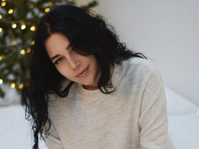 LiyaFrench with long black wavy hair, wearing a grey sweater, leaning forward with a gentle smile, soft brown eyes, and a subtle, sexy, cute, and attractive look.