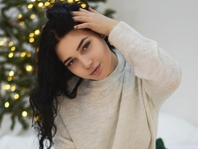 LiyaFrench, con lunghi capelli neri, indossa un maglione grigio, con una mano tra i capelli, guarda leggermente verso il basso, con un sorriso dolce. In secondo piano, un albero di Natale con luci gialle, leggermente sfocato..