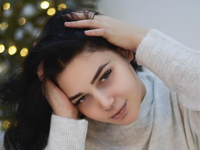 LiyaFrench, com cabelo preto, as mãos no cabelo, vestindo um suéter cinza, tem um rosto fofo e ligeiramente irritado, pele macia e olhos castanhos. Sem seios, sem bumbum ou vulva visíveis..