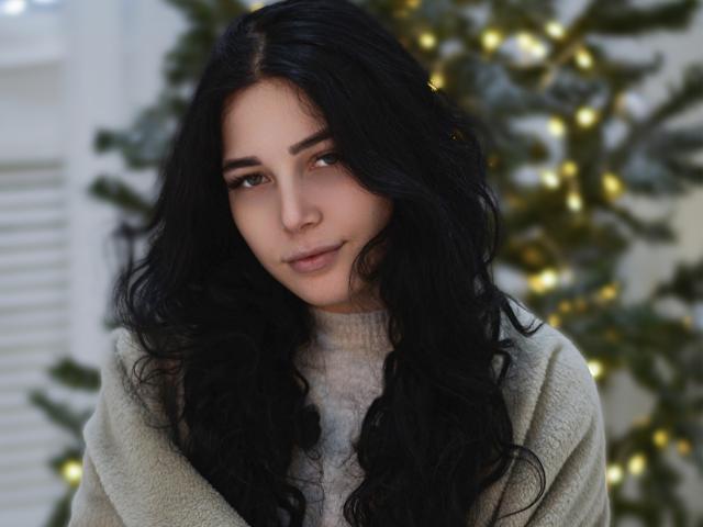 LiyaFrench with long, wavy black hair, light skin, and subtle makeup, wearing a beige sweater, looking slightly to the side with a gentle smile. Christmas tree in the background.