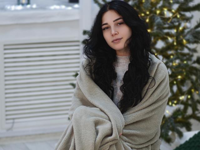 LiyaFrench with long black wavy hair, green eyes, and a soft smile, wrapped in a beige blanket, wearing a gray sweater, sitting in front of a Christmas tree.