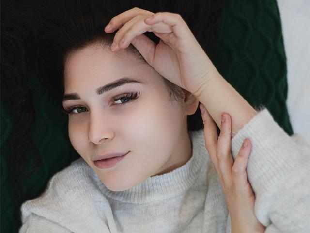 LiyaFrench, con cabello rizado oscuro, ojos verdes y piel clara, vestida con un suéter blanco, acostada con las manos tocando su cabeza y cuello, mirando a la cámara..
