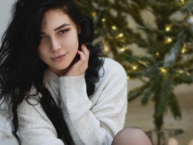 LiyaFrench, com longos cabelos pretos, vestindo um suéter branco, sentada em frente a uma árvore de Natal com luzes amarelas, com a mão tocando sua bochecha, olhando para a câmera e sorrindo..