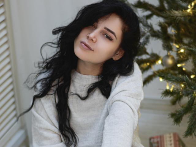 LiyaFrench with wavy black hair, light skin, and a nose ring, wearing a white sweater, leaning against a decorated Christmas tree. Her tits are slightly visible under the sweater.