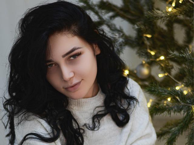 Liya, con cabello negro ondulado, piel clara y ojos verdes, vestida con un suéter blanco, está de pie junto a un árbol de Navidad con luces amarillas. Está sonriendo suavemente y mirando a la cámara..