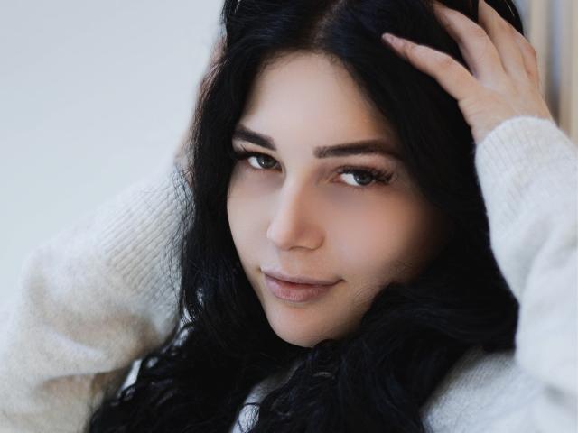LiyaFrench has long black hair, a cute face, and is looking at the camera with a slight smile. She's wearing a white knit sweater, touching her head.
