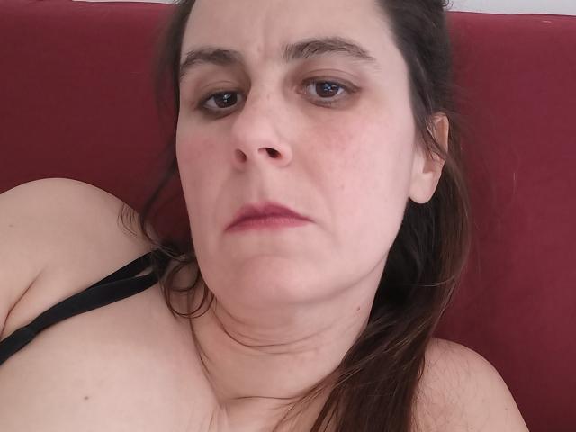 FetishFeminity heeft een serieus, licht gezwollen gezicht met bruine ogen, lichte huid en bruin haar. Ze draagt een zwarte band over haar schouder, waardoor haar blote schouder en bovenste borst te zien zijn..