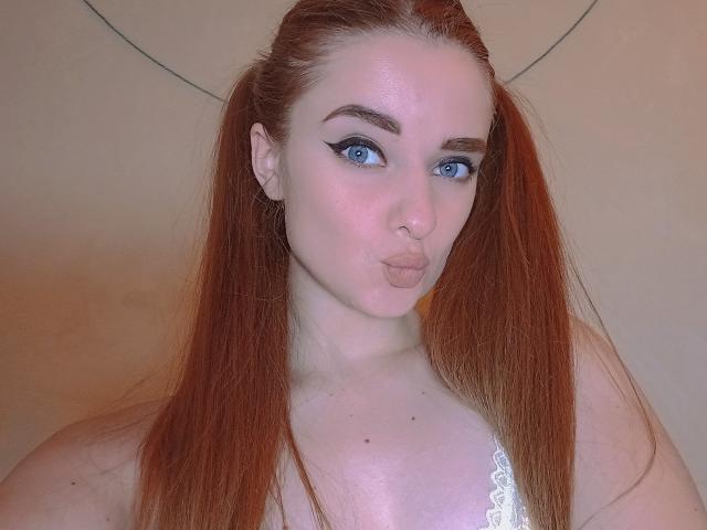 KariMillie with puffy red hair in pigtails, blue eyes, and white lace top, making a pouty face. Her tits are small and her ass is not visible.