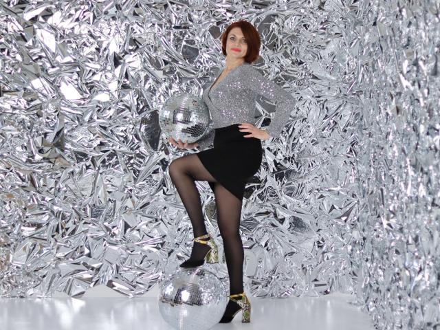 LisaTreds with red hair, sparkling gray top, black skirt, sheer black stockings, and gold heels, standing on a silver ball, holding another ball, looking confident.
