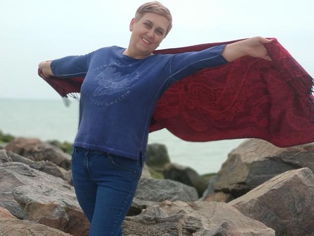 MadamMargo with short brown hair, blue sweater, and red patterned scarf, standing on rocky shore, arms out, smiling, wearing blue jeans, holding scarf open.