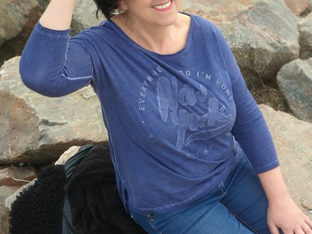 MadamMargo with medium tits, wearing a blue shirt and jeans, smiling, black bag on rock, hand in hair, outdoor setting.