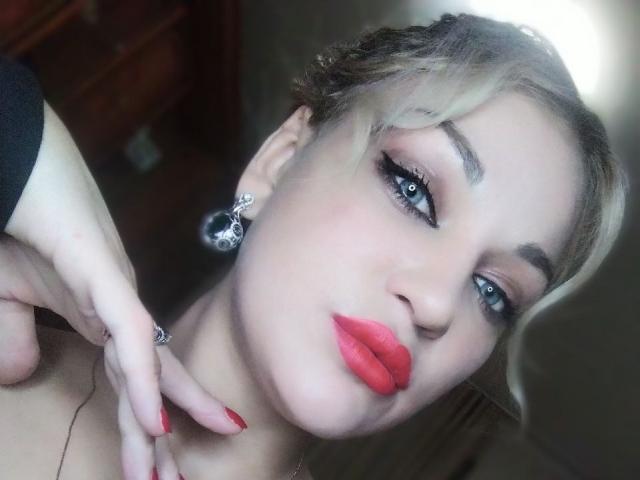 EvaAlferash, con cabello rubio, ojos azules, lápiz labial rojo y delineador de ojos negro, lleva aretes negros y un anillo, y está tocándose el cuello con los dedos pintados de rojo..