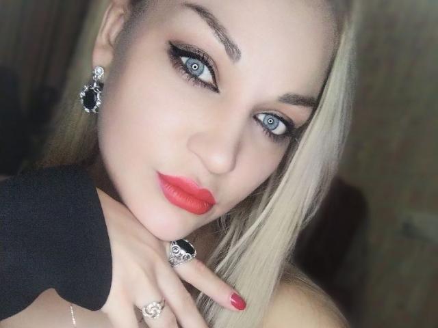 EvaAlferash with blonde hair, blue eyes, red lipstick, black earrings, black ring, black necklace, and red nail polish, looking at the camera with a slight smile.