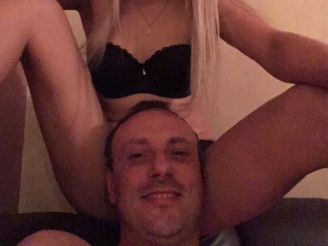 EvaAndAnto with blonde hair, wearing black lace bra, straddles a smiling bald man with a mustache, her legs on either side of his head, in a dimly lit room.