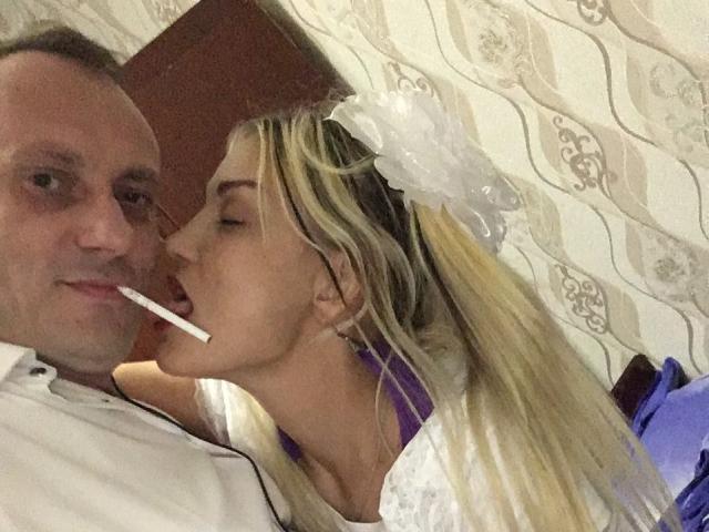 EvaAndAnto, com cabelo loiro e uma coroa de flores brancas, suga um cigarro de um homem careca vestindo uma camisa branca, com sua blusa roxa visível, olhos fechados, em um quarto..
