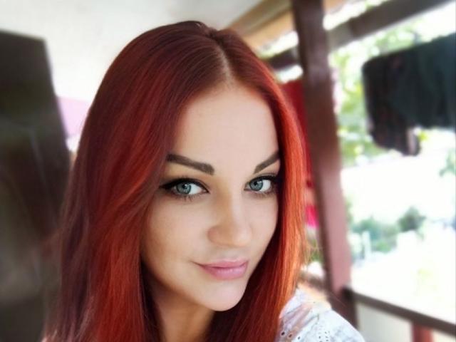 EvaAndAnto ha i capelli rossi, gli occhi azzurri e un sorriso carino, con i seni leggermente visibili sotto una camicia bianca, e guarda direttamente la fotocamera..
