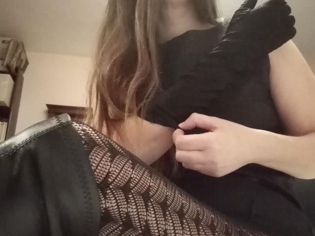 Femminilità sensuale con lunghi capelli castani, che indossa un abito nero, sollevandolo per rivelare calze nere con motivi in pizzo e pelle nera. La sua mano è sull'abito..