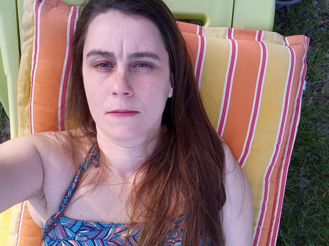 Een vrouw met een fetisjistische uitstraling, met bruin haar, lichte huid en kleine borsten, draagt een kleurrijke bikini-top met een patroon en ligt op een oranje en gele gestreepte stoel, met een licht verveelde blik..