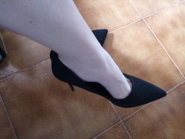 La jambe pâle de FetishFeminity, chaussée d'une haute talon pointu noire, descend un escalier, montrant son pied à l'intérieur de la chaussure. Un sol carrelé est visible en arrière-plan..