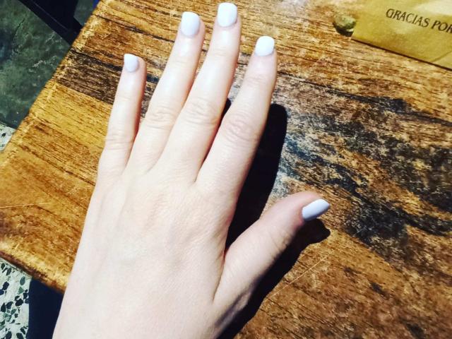 La main de FetishFeminity, avec des ongles peints en blanc, repose sur une surface en bois, révélant un teint clair et lisse, ainsi que des ongles soigneusement coupés. Le bois présente un grain et une texture visibles..