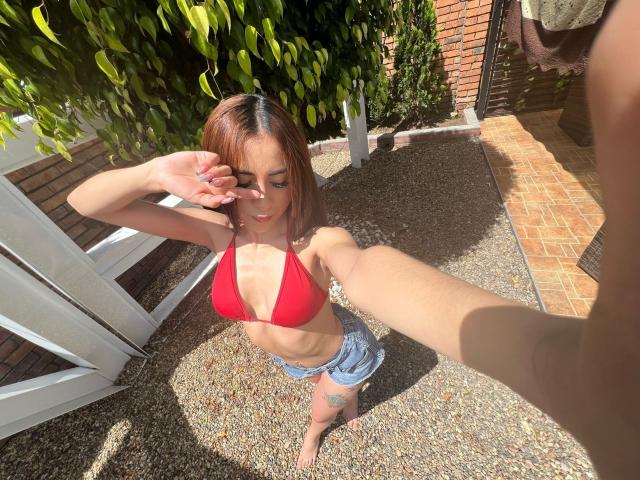 AnyaJay with small red boobs, wearing a red bikini top and blue denim shorts, taking a selfie, her hand covering her eye, standing on gravel, sunny, tattoo on her right thigh.