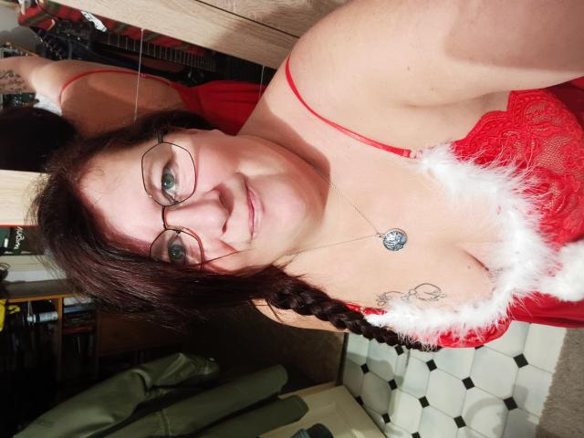 InstantShow with red lace dress, white fur trim, braids, glasses, and a necklace, smiling at the camera, showing her tits and tattoo on her right shoulder.