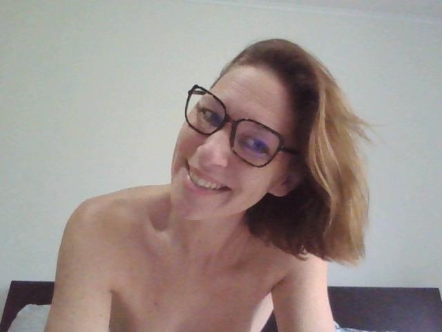 ApoloniaRed, topless with glasses, smiling at the camera, brown hair, small tits, fair skin, leaning forward, in a bedroom.