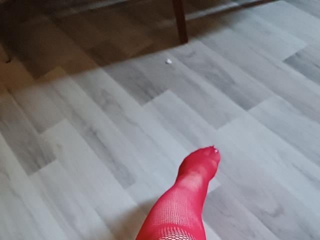 PenJanis' roter Fuß, bedeckt mit Fischernetz, mit einem kleinen Loch in der Nähe der Zehen, steht auf einem hellen Holzboden. Das Foto wurde aus einem niedrigeren Winkel aufgenommen und zeigt den Fuß..