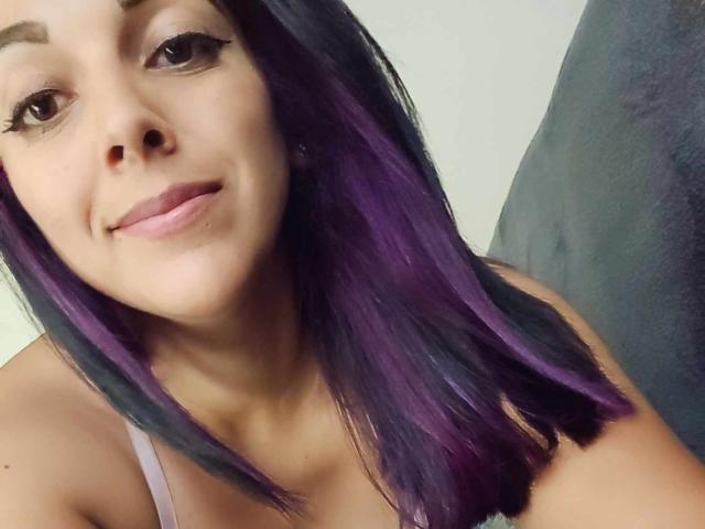 PrettyEllie with purple and black hair, smiling slightly, has her tits barely covered by a pink tank top, brown eyes, and a cute face. Her ass isn't visible.