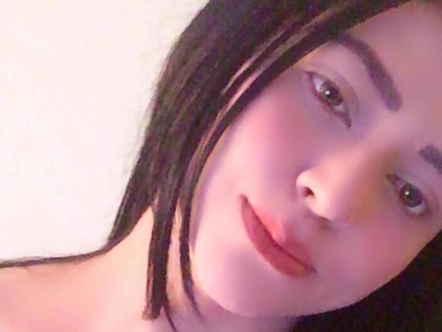 O rosto de Katirra, com pele rosada, olhos castanhos e cabelo preto e liso, lábios levemente entreabertos e um sorriso discreto. Ela está sem blusa, mostrando seus ombros nus. Iluminação suave e rosada..