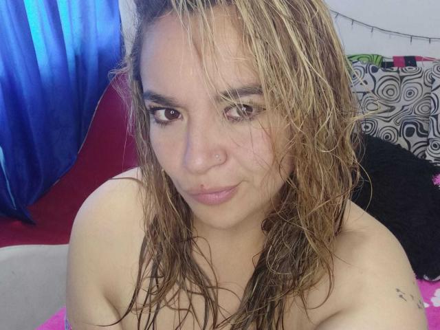 NaugthyCurvy with wet, blonde hair, brown eyes, and a small nose piercing, looking at the camera with a slight smile, showing bare shoulders and a small tattoo on her arm.