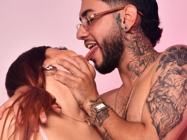 AdharaAndSimon, a red-haired chick with glasses, gets her facehanded by Simon, a tattooed dude with glasses, sticking his tongue down her neck. He's licking her face.