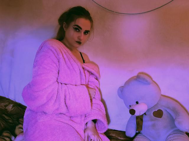 KariMillie met een roze, pluizige badjas, donkere lipstick en een witte teddybeer, zittend op het bed, kijkend naar de camera, met roze en paarse verlichting, waarbij ze haar borsten licht bedekt met de badjas..