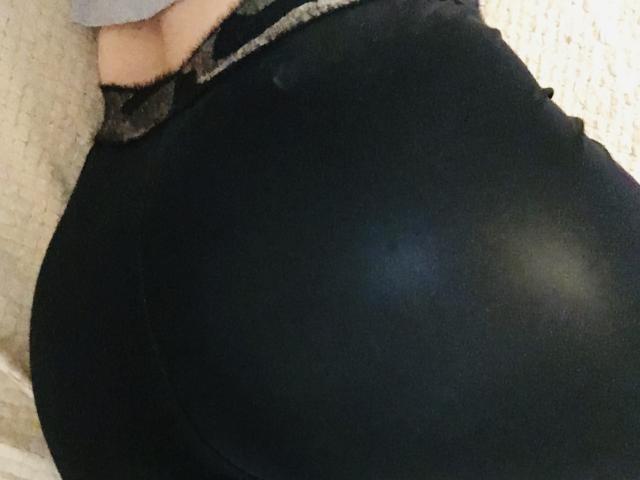 Le postérieur noir et brillant de Barbarica est bien visible dans le cadre, avec une petite partie de sa culotte, bordée de dentelle, qui est visible en haut. Un tissu doux et texturé se trouve en arrière-plan..