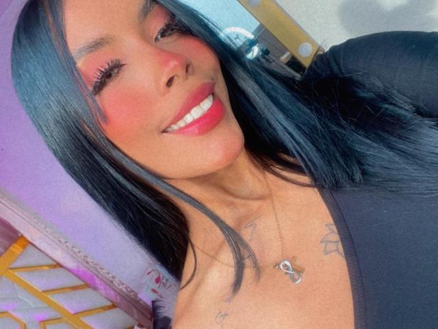 FiorellaBossh hat glänzendes, schwarzes Haar, rosigen Lippen und ein breites Lächeln mit pinkem Lippenstift. Sie trägt ein schwarzes Oberteil und eine Halskette mit einem Herzanhänger und zeigt Tattoos..