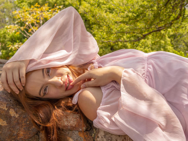 MadeleineBlack lying on a rock, wearing a pink, off-shoulder dress, with her hand touching her red lips, smiling, green eyes, red hair, sunlight, and a lush green background.
