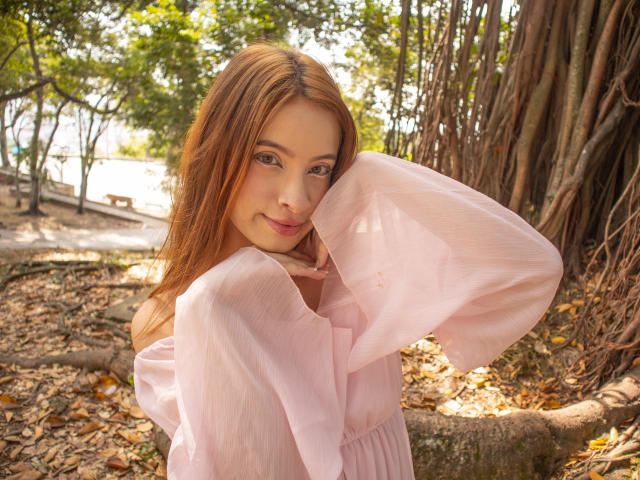 Madeleine, con cabello largo y castaño, vestido rosa pálido con un hombro descubierto, una sonrisa suave y un rostro adorable, sosteniendo su barbilla, de pie junto a un árbol con raíces gruesas y hojas verdes..